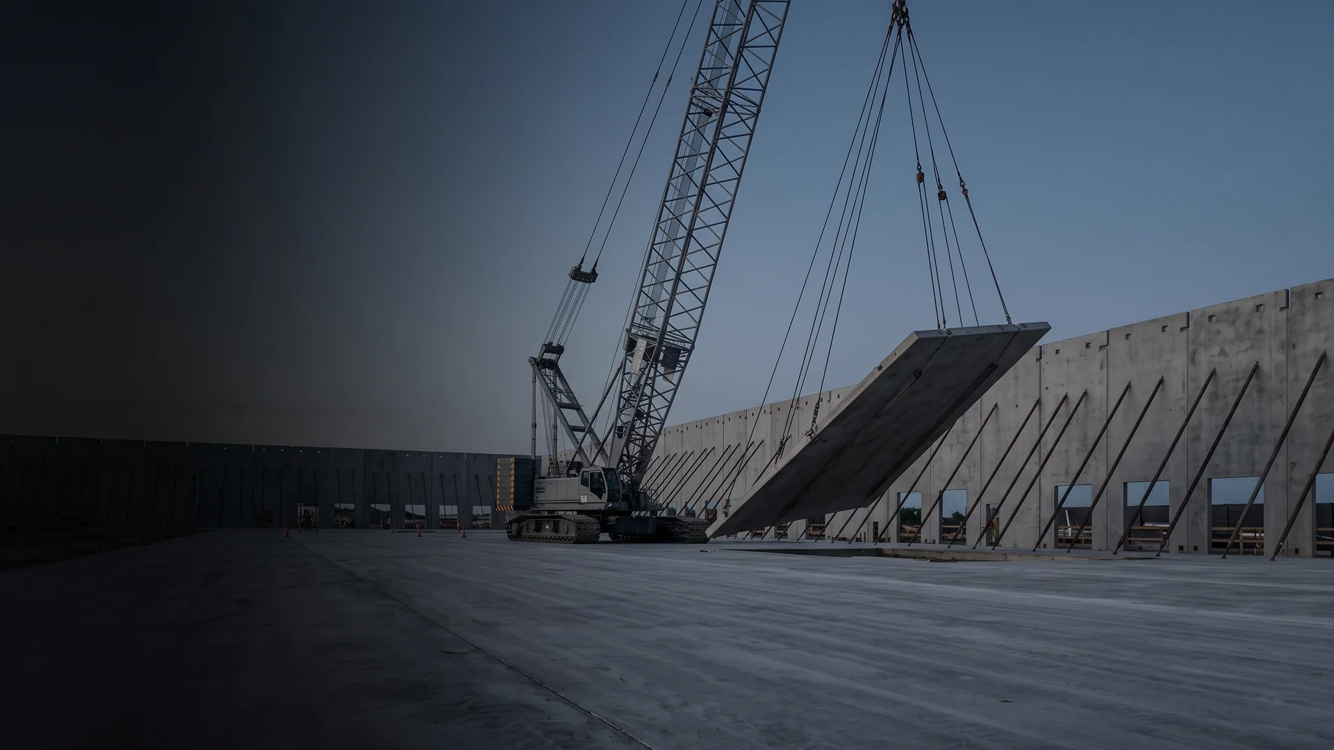 Crane lifting a tilt-wall concrete panel upright at dawn on an industrial construction site